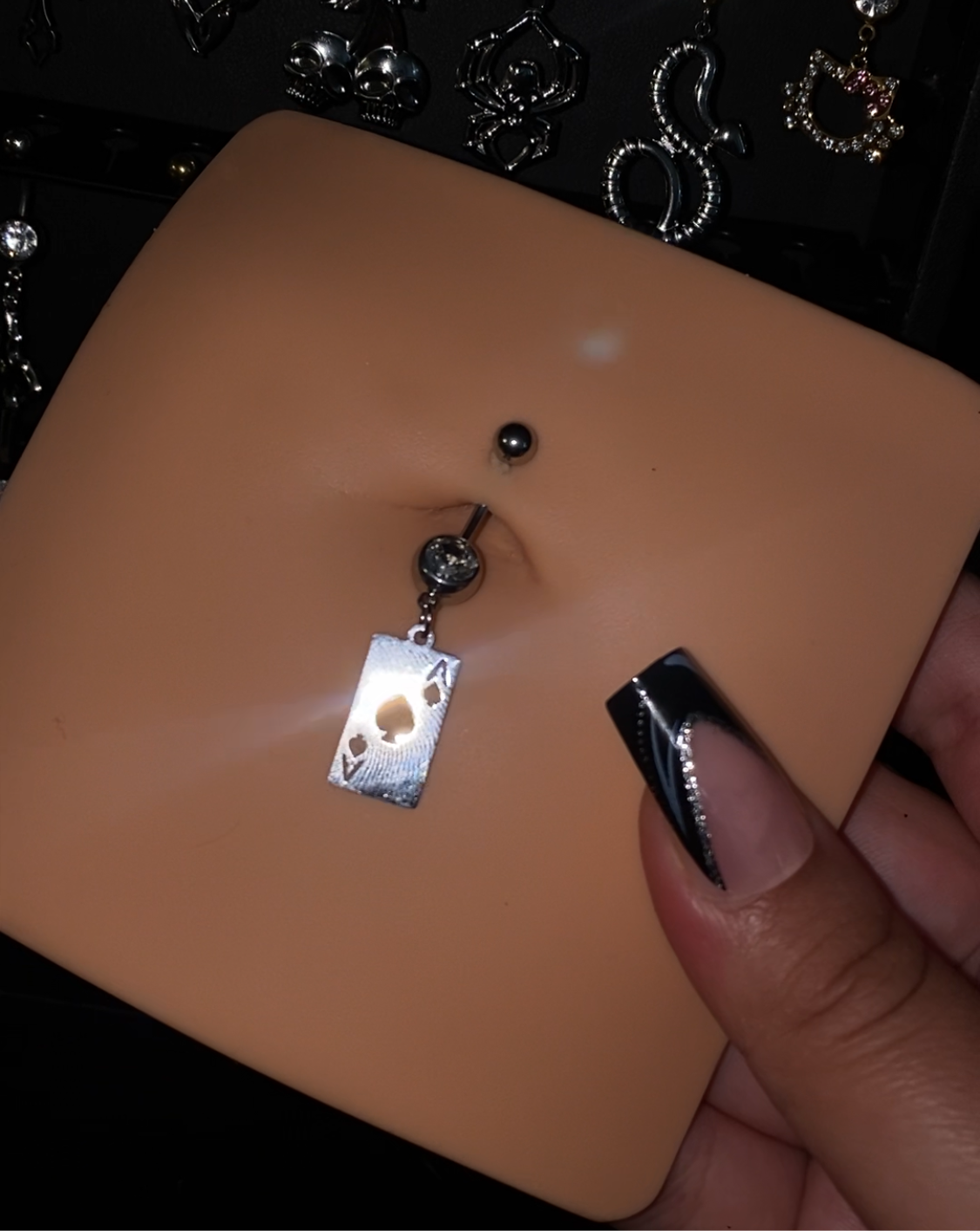 Ace Belly Button Piercing, featuring a stainless steel charm in the shape of an Ace card, with a 14g externally threaded 316L Stainless Steel/Titanium bar, available in 10mm, 12mm, and 14mm lengths, displayed on a mannequin with a background of decorative piercings.