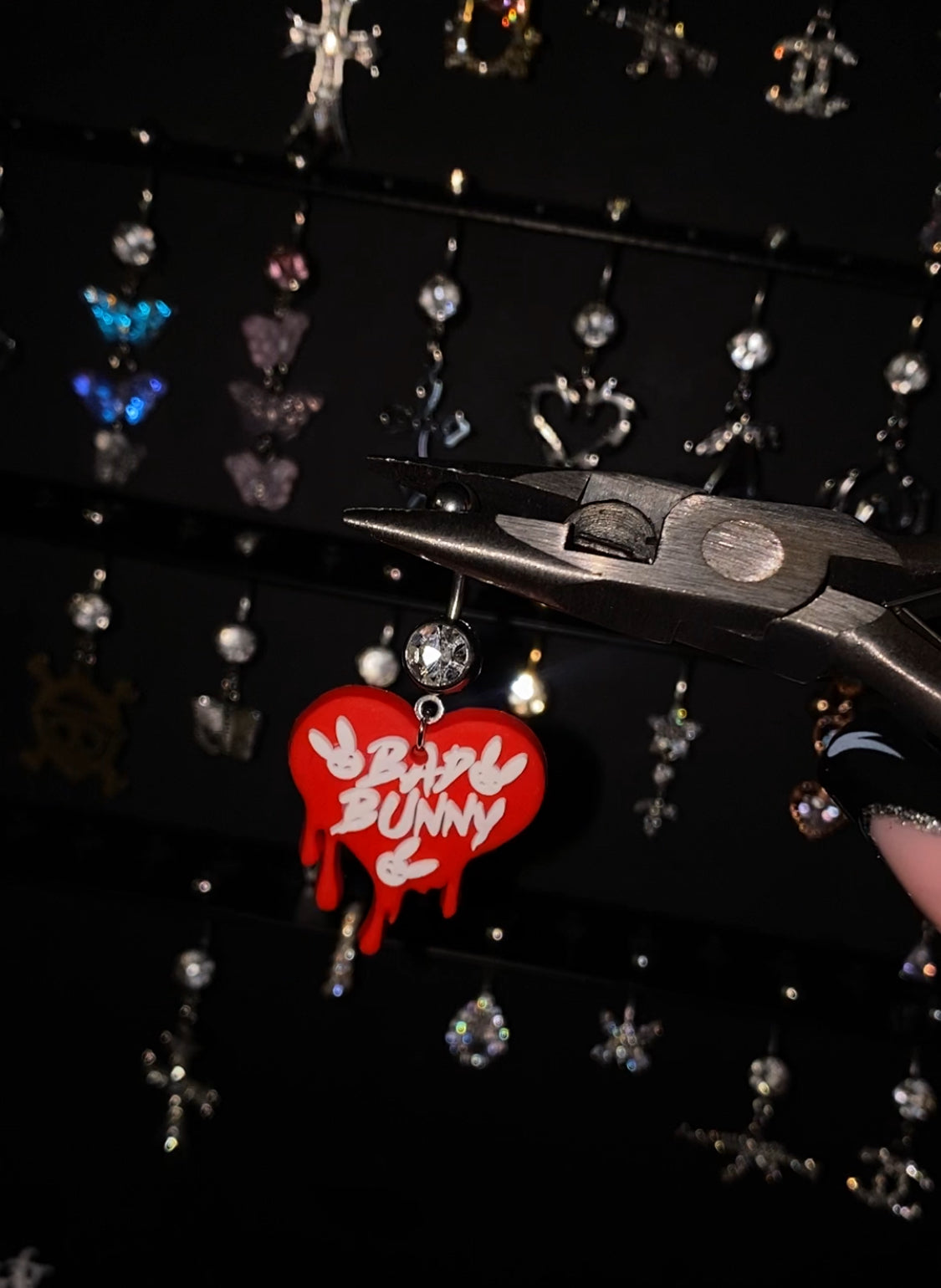 Close-up of the "Bad Bunny Belly Button Ring" held by pliers against a backdrop of assorted jewelry, featuring a red, heart-shaped charm with "Bad Bunny" text, made from 316L Stainless Steel and ASTM F-136 Titanium, waterproof, tarnish-free, 14g thickness, and available in lengths of 10mm, 12mm, and 14mm.