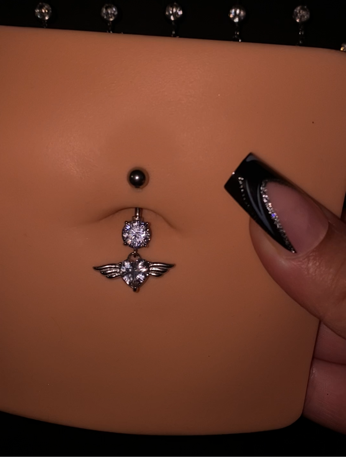 "Angel Belly Button Piercing with stainless steel and titanium bar, featuring a sparkling charm with wings and a gem, showcased on a mannequin, waterproof and tarnish-free, available in various lengths and thicknesses."